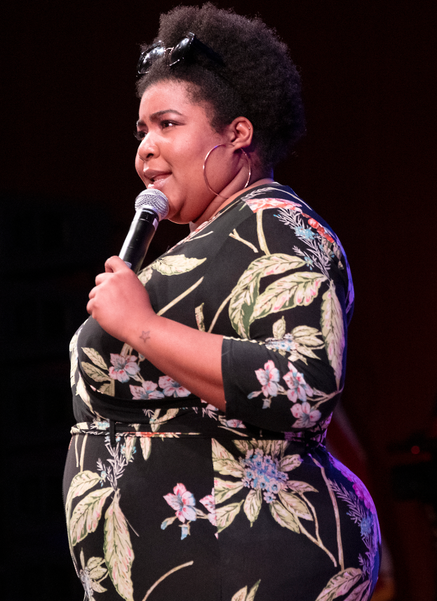 Eine Frau in einem schwarzen Kleid steht selbstbewusst auf einer B├╝hne, h├Ąlt ein Mikrofon in der Hand und ist bereit, aufzutreten.