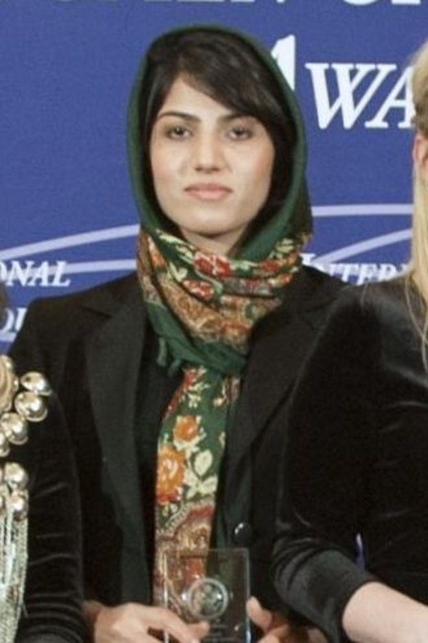 Malala Yousafzai, eine Nobelpreisträgerin für den Frieden, steht in der Mitte und trägt ein schwarzes Kleid und einen Schal, hält eine Auszeichnung und ist von zwei Frauen in schwarzen Kleidern flankiert, mit einem blauen Banner im Hintergrund.