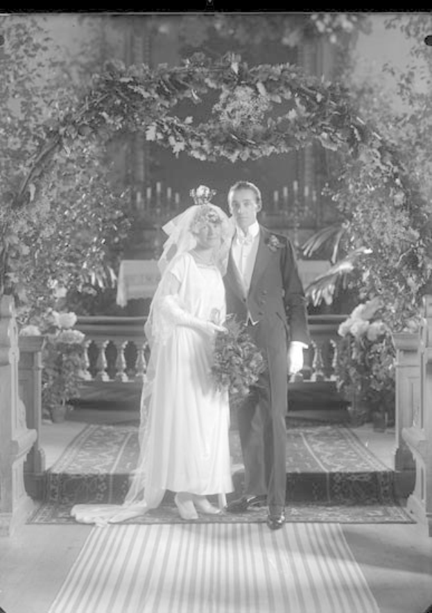 Schwarz-weiß-Foto eines Mannes und einer Frau vor einem Blumenschmuckbogen, die Frau hält einen Strauß Blumen, mit einem Teppichboden.