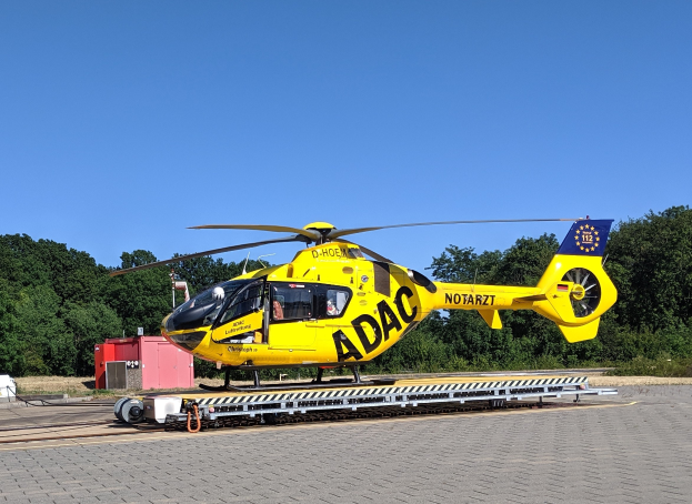 Gelbes Helikopter auf einem Flughafen-Asphalt mit umliegenden Bäumen und einem klaren blauen Himmel, wahrscheinlich ein Luftrettungshelikopter in Deutschland.