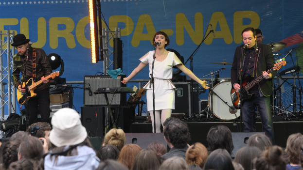 Eine Frau in einem weißen Kleid singt auf der Bühne bei einem Konzert, flankiert von zwei Gitarristen, mit einer Menge davor und Lautsprechern im Hintergrund.