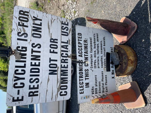 Ein Schild mit der Aufschrift "E-Bike-Fahren nur für Anwohner" steht an der Seite einer Straße, umgeben von Verkehrskegeln und anderen Gegenständen, mit einem Fahrzeug, Bäumen und einem klaren blauen Himmel im Hintergrund.