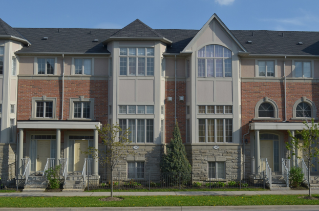 Reihe von Stadthäusern mit Fenstern, Türen, Säulen, Geländern, Treppen, Pflanzen, Bäumen, Gras, einer Straße im Vordergrund und Himmel im Hintergrund.