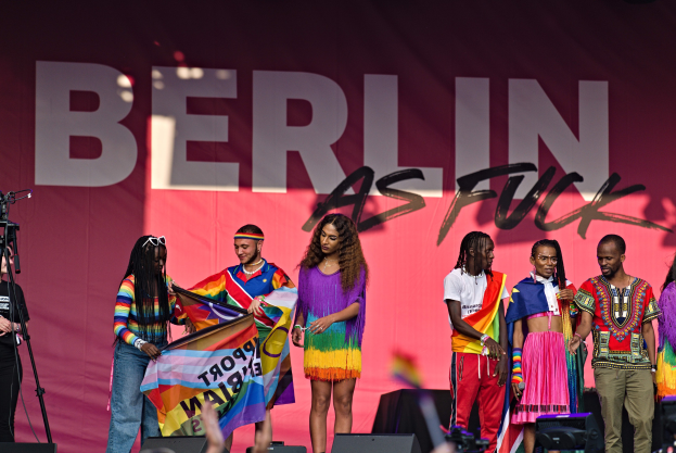 Eine Gruppe von Menschen auf einer Bühne mit einer Frau, die eine Flagge in der Mitte hält, einer Person, die eine Kamera auf einem Stativ links bedient, und einem Banner im Hintergrund mit der Aufschrift "Berlin als Fuck 2019".