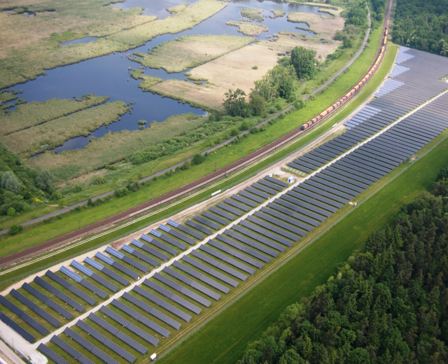 Luftaufnahme eines Solarparks mit Panelen, umgeben von Bäumen, Gras, Wasser und einer nahen Bahnlinie.