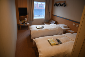 Ein Hotelzimmer mit zwei Betten, einem Wandfernseher, einem Fenster mit Vorhängen, Bügeln auf der rechten Seite und einem Tisch mit verschiedenen Gegenständen vor den Betten.