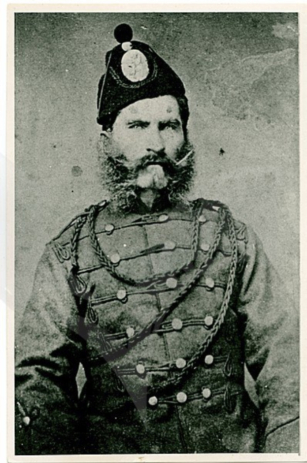 Altes Schwarz-Weiß-Foto eines bärtigen Mannes in militärischer Uniform mit Schirmmütze, der einen Gürtel mit einer Schnalle trägt und direkt in die Kamera blickt.