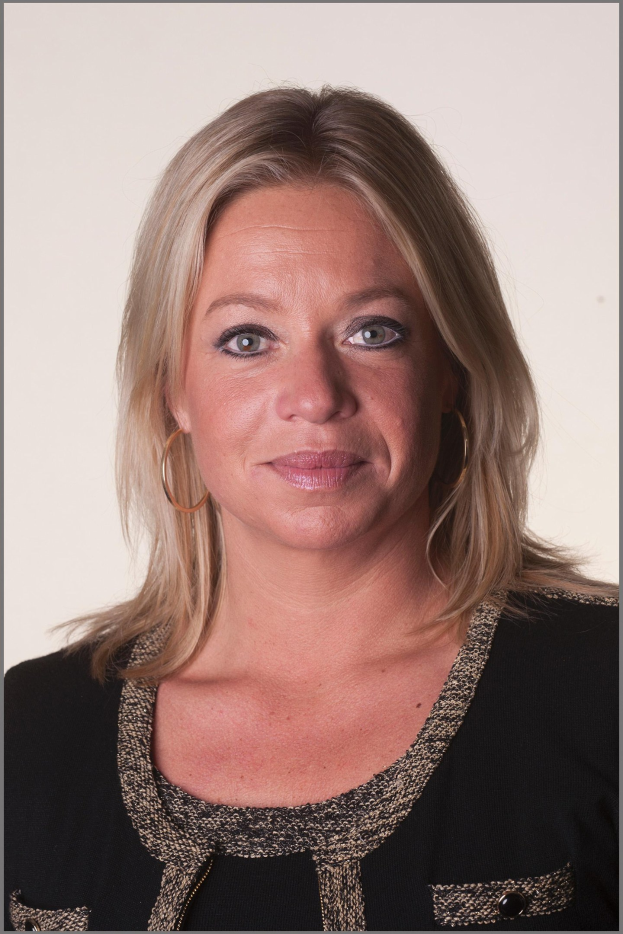 Eine Frau mit blondem Haar in einem ordentlichen Dutt, die ein schwarzes Kleid und goldene Kreolen trägt und einen warmen und selbstbewussten Ausdruck hat.