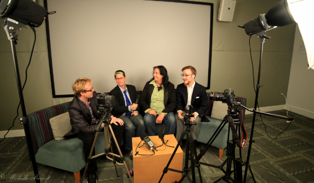 Eine Gruppe von drei Personen sitzt auf einem Sofa und gibt ein Interview, wobei eine Person links in einem schwarzen Mantel und Hose sitzt, eine Person in der Mitte einen blauen Schlips trägt und ein Mikrofon hält und eine Person rechts lächelt. Hinter ihnen befindet sich ein Projektionsschirm, ein Mikrofonrecorder auf dem Tisch in der Mitte und Kameras mit großen Stativen auf beiden Seiten.