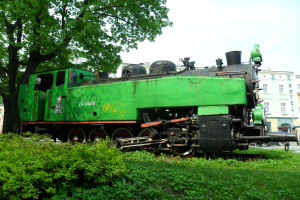 Grüner Zugmotor auf einer saftig grünen Wiese mit Pflanzen und Bäumen, Gebäuden, Fahrzeugen und einem klaren blauen Himmel im Hintergrund.
