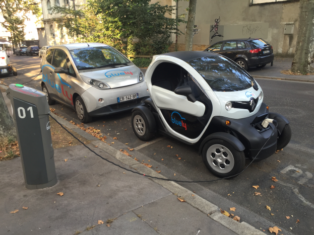 Zwei Elektroautos nebeneinander auf einer Straße geparkt, an eine Ladesäule angeschlossen, mit Bäumen und Gebäuden mit Fenstern im Hintergrund.