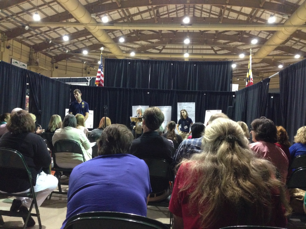 Eine Gruppe von Menschen, die auf Stühlen sitzen, mit einer Person, die an einem Rednerpult steht, in einem Raum mit Transparenten, blauen Vorhängen, Flaggen und Deckenleuchten.