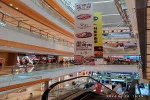 Einblicke in ein Einkaufszentrum mit einer Rolltreppe in der Mitte, Menschen, die herumlaufen, einige mit Handtaschen, und Schilder, die von der Decke hängen, mit Geschäften im Hintergrund.