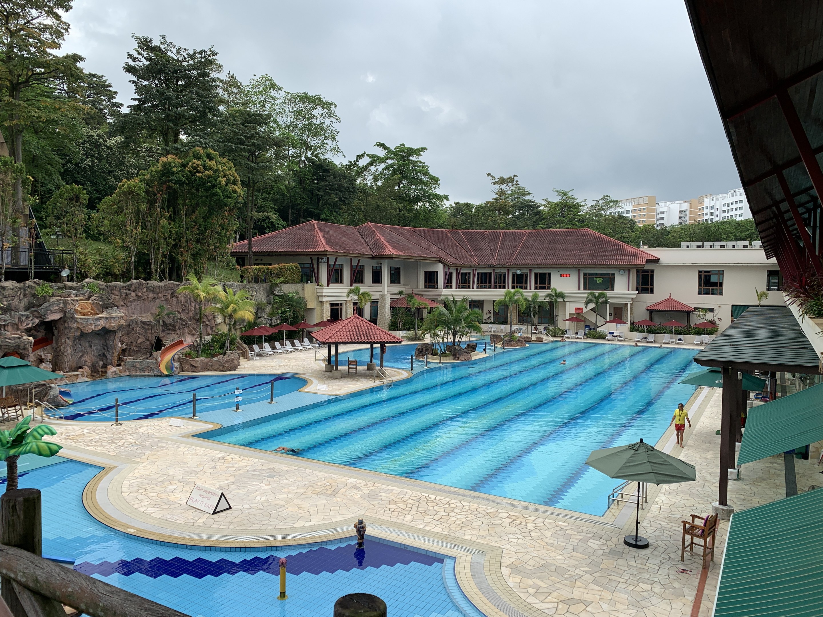 Großes Schwimmbad im Ritz-Carlton, Kota Kinabalu, mit Menschen, Sonnenschirmen, Stühlen, Tischen, Pflanzen, Bäumen, Felsen, Gebäuden, einer Hütte rechts und einem sichtbaren Himmel im Hintergrund.