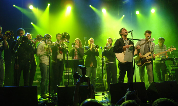 Eine Gruppe von Musikern auf der Bühne mit Mikrofonen, Scheinwerfern und einer Kamera, während das Publikum zusieht.