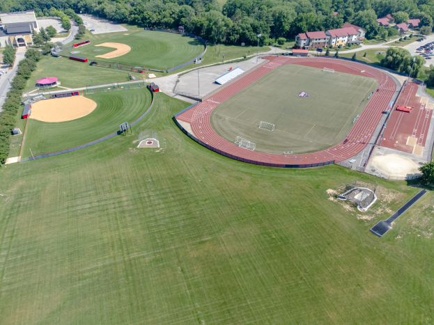Eine Luftaufnahme eines Baseballfelds mit grünem Rasen, Bäumen, Gebäuden und Fahrzeugen auf einer nahen Straße.