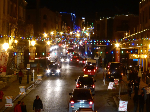 Eine belebte Straßenszene mit verschiedenen Fahrzeugen, darunter Autos, Lastwagen und Lieferwagen, Gebäuden auf beiden Seiten, Fußgängern und Passanten, Bannern, Werbetafeln, Laternenmasten, Dekorationslichtern und Bäumen.
