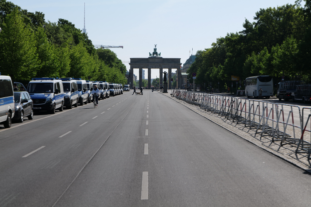 Eine Reihe von Polizeiwagen auf einer Straße vor dem Brandenburger Tor in Berlin geparkt, mit Menschen auf Fahrrädern und in der Nähe Stehenden, Barrieren, Bäumen und einem Bogen mit Statuen im Hintergrund.