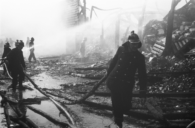 Schwarzes und weißes Bild von Feuerwehrmännern in Helmen, die durch die Tr√∞mmer eines zerstörten Gebäudes gehen und Rohre tragen.