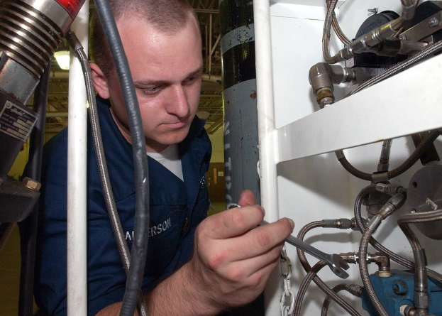 Techniker in blauer Uniform, der Maschine inspiziert oder justiert, umgeben von Industrieausrüstung, Rohren und Beleuchtung.
