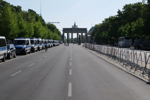 Lange Reihe von Polizeiwagen auf der Straße vor dem Brandenburger Tor in Berlin, Deutschland geparkt, mit Menschen auf Fahrrädern und auf der Straße, Barrieren, Bäume, ein Tor mit Statuen im Hintergrund und sichtbarer Himmel.