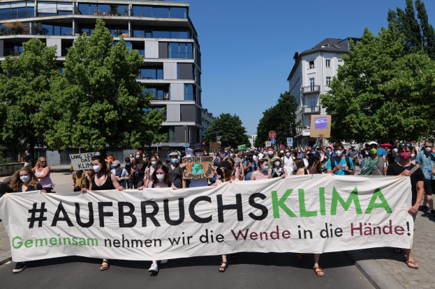 Eine Gruppe von Menschen mit Masken, die eine Fahne mit der Aufschrift 'Aufbruchsklima' vor einem Gebäude mit Bäumen und einem klaren Himmel halten.