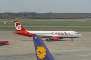Ein Air-Berlin-Airbus A320-200 auf der Rollbahn am Frankfurter Flughafen, umgeben von Fahrzeugen auf der linken Seite und Gras am Boden. Im Hintergrund sind Pole, Bäume und ein klarer blauer Himmel zu sehen.