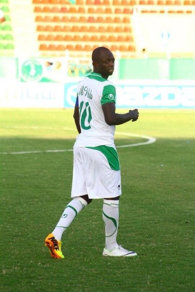 Ein Fußballspieler in grüner und weißer Uniform feiert auf einem Feld umgeben von Werbetafeln und Stühlen, wahrscheinlich nach dem Gewinn des Afrika-Cups.