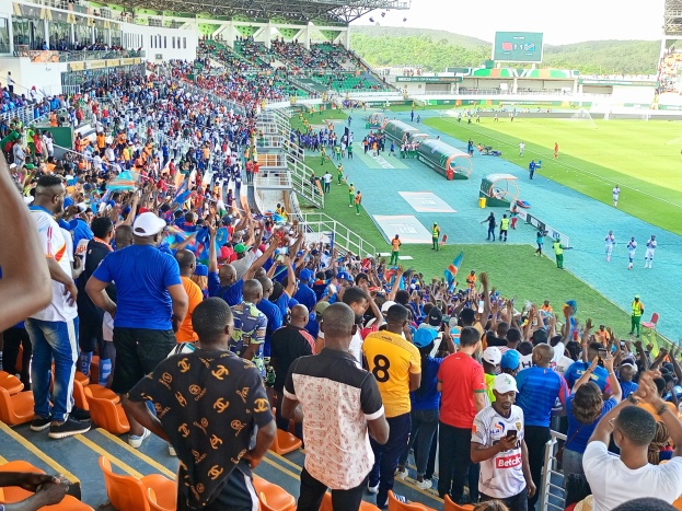 Eine große Menschenmenge versammelt sich vor einem eingezäunten Fußballfeld, wobei einige auf Stühlen sitzen und andere stehen, unter einem klaren blauen Himmel mit Hügeln im Hintergrund und harren der 2019 African Cup of Nations-Partie.