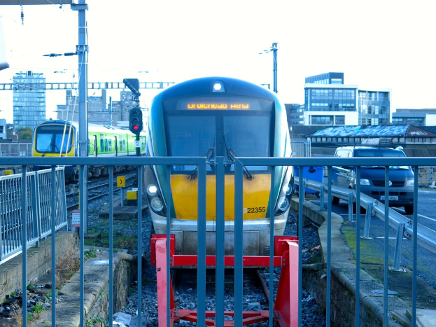 Zwei Züge auf parallelen Gleisen mit einem Zaun im Vordergrund, Strommasten einschließlich einem mit einer Signalleuchte, ein Auto auf einer Straße rechts, Gebäude im Hintergrund und Himmel darüber.