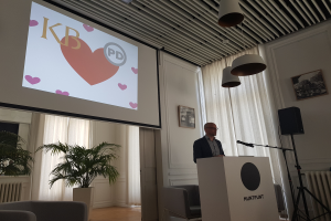 Ein Mann steht an einem Rednerpult mit einem Mikrofon und hält eine Präsentation vor einem Projektionsschirm, mit Stühlen, einer Pflanze, Fotorahmen, Vorhängen und Deckenlampen im Hintergrund.