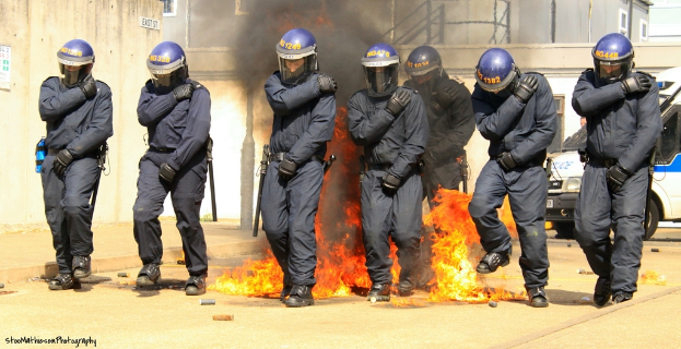 Menschen in Helmen stehen vor einem Feuer mit Gegenständen auf dem Boden, Gebäude im Hintergrund, ein Fahrzeug, ein Plakat und eine Tafel an der linken Wand und Text unten.