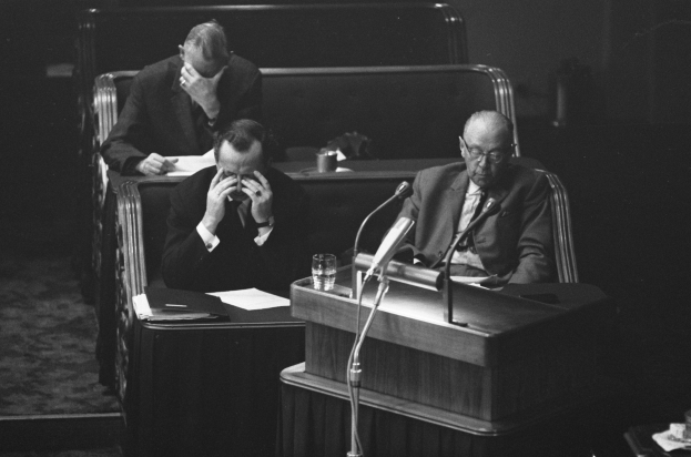 Zwei Männer sitzen an einem Podium mit Mikrofonen, umgeben von Tischen mit Papieren und anderen Gegenständen, in eine Diskussion vertieft, dargestellt in Schwarz-Weiß.