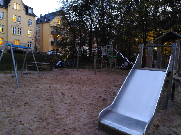 Ein Spielplatz mit einer Rutsche in der Mitte, umgeben von Sand, Schaukeln, Pfosten und anderen Geräten, mit Bäumen, Gebäuden, Fahrzeugen und einem klaren blauen Himmel im Hintergrund.