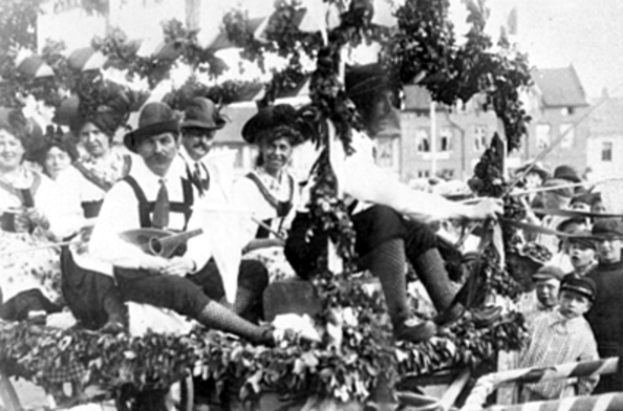 Eine Gruppe von Menschen in traditioneller bayerischer Tracht fährt auf einem mit Blättern geschmückten Festwagen mit, einige spielen Musikinstrumente, mit Gebäuden und einem klaren Himmel im Hintergrund, dargestellt in Schwarz-Weiß.