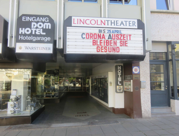Außenansicht des Lincoln Theaters in Berlin, Deutschland, mit Glasfenstern und -türen sowie einer Tafel mit Text und einem Innenblick, der eine belebte Stadtlandschaft mit verschiedenen Gegenständen zeigt.