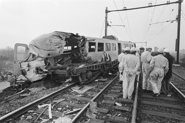 Schwarze und weiße Szene eines Zugunglücks auf den Schienen mit Menschen in Helmen in der Nähe, Strommästen, Bäumen, Gebäuden und einem klaren Himmel im Hintergrund.