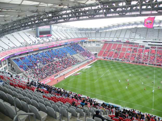 Ein großes Stadion voller Zuschauer bei einem Fußballspiel, mit Spielern im Einsatz auf dem Feld und einem Bildschirm, einer Hütte und Bannern oben.