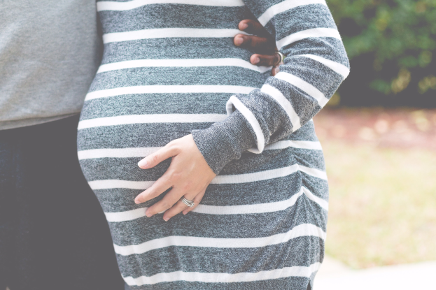 Schwangere Paare in Umstandskleidung, die zusammenstehen und den Bauch berühren, umgeben von Bäumen.