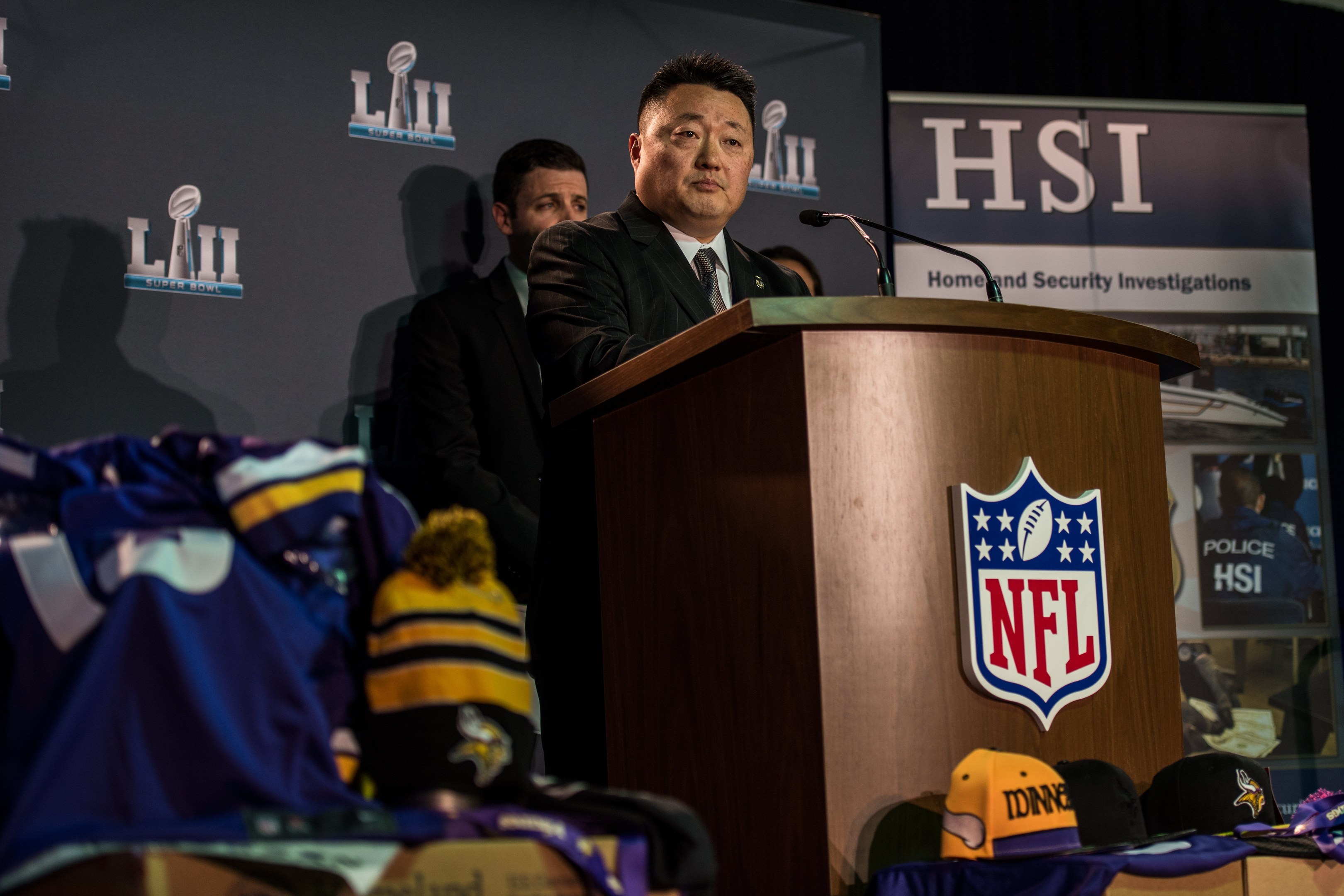 Ein Mann in einem schwarzen Blazer, einem wei├čem Hemd und einer schwarzen Krawatte spricht an einem Podium mit einem Mikrofon und Logo, gewandt einer Menge mit einem "Super Bowl LII"-Banner dahinter, waehrend blaue und gelbe Merchandising-Artikel links von ihm angezeigt werden.