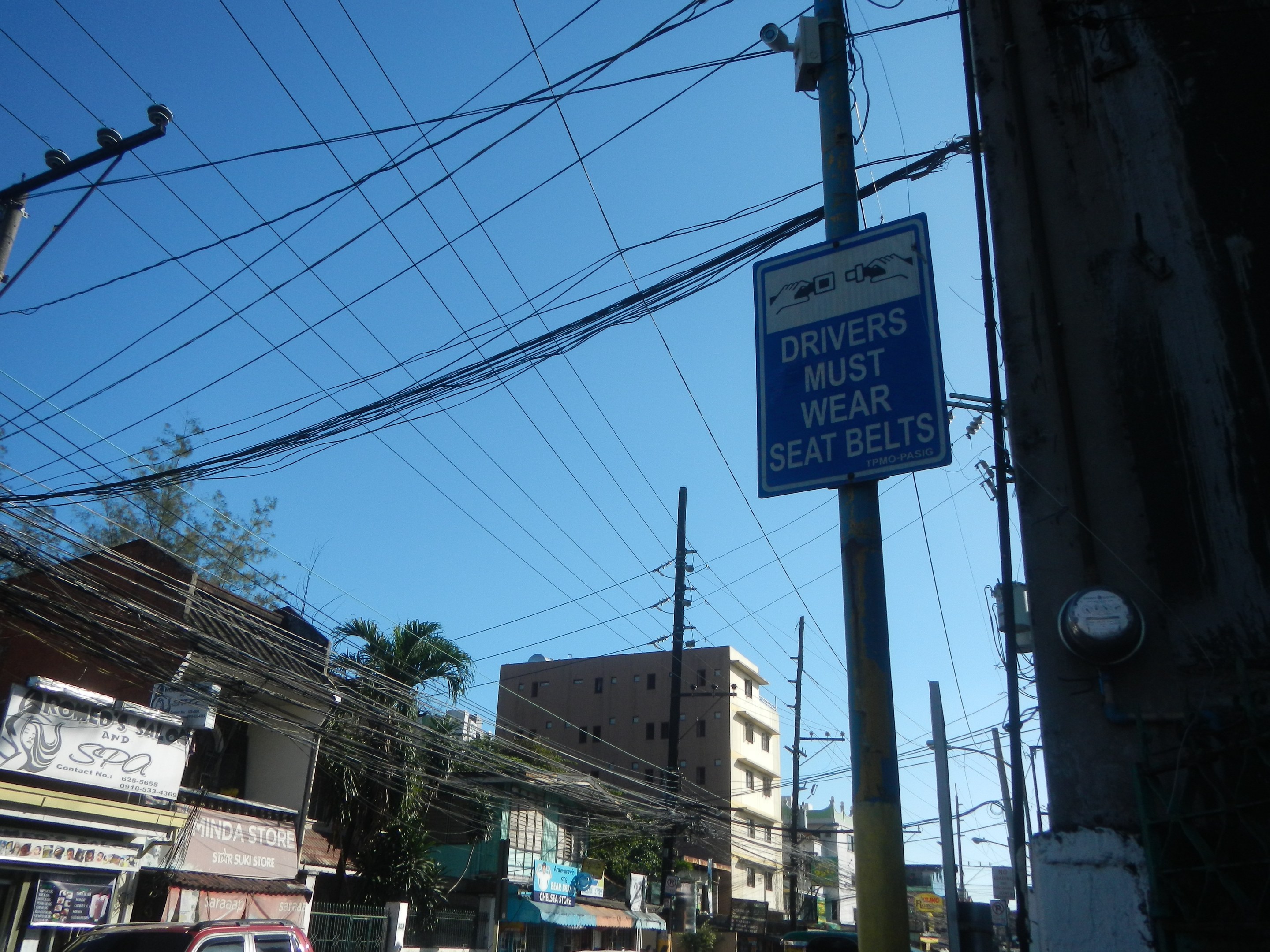 Stadtstraße mit fahrenden Autos, Strommasten mit Kabeln, Gebäuden, Bäumen und Namensschildern, mit einem "Fahrer müssen den Sicherheitsgurt anlegen"-Schild an einem Strommast.