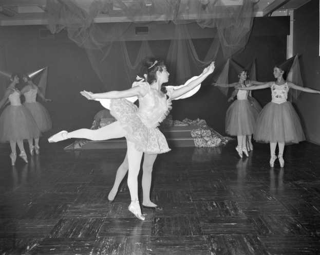 Eine Gruppe junger Balletttänzerinnen in Tutus und Spitzenschuhen tanzt auf einer Bühne mit einer Wand im Hintergrund und einer Decke mit Lichtern oben, dargestellt in Schwarz-Weiß.