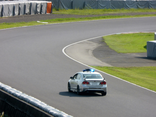 Polizeiauto auf einer Rennstrecke mit Gras auf beiden Seiten, umgeben von Menschen, Zelten und anderen Gegenständen im Hintergrund.