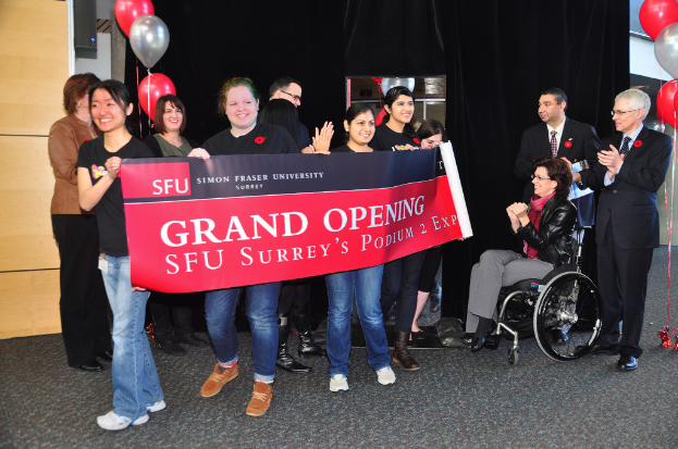 Eine Gruppe von Frauen hält ein Banner hoch und bewirbt ein Event, während zwei Männer daneben klatschen und eine sitzende, aufgeregte Frau im Rollstuhl zu sehen ist, vor einem schwarzen Vorhang und Ballons.