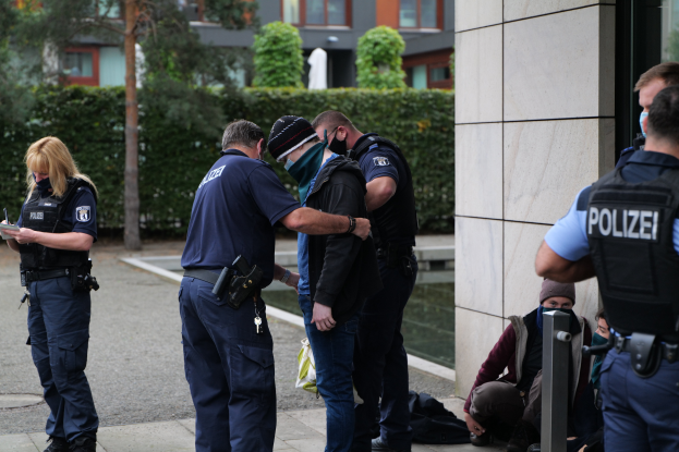 Eine Gruppe von Polizisten steht vor einem Gebäude, eine Frau hält ein Buch und einen Stift in der Hand, während mehrere Personen stehen und sitzen und Pflanzen, Bäume und Gebäude im Hintergrund zu sehen sind.