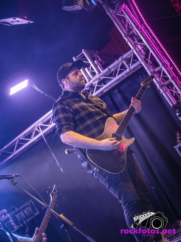 Ein Mann in einer Mütze spielt Gitarre auf der Bühne eines Konzerts, umgeben von Mikrofonen, Lichtern, Trägern und anderen Musikinstrumenten, mit Text unten lesend "Bruno Mars - Rockfotos Net".