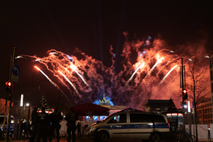 Ein Polizeiwagen steht vor einer Menschenmenge, mit beleuchteten Gebäuden, Bäumen, Polen, Verkehrszeichen, Schildern und Zelten im Hintergrund, während bunte Feuerwerke den Himmel erhellen.