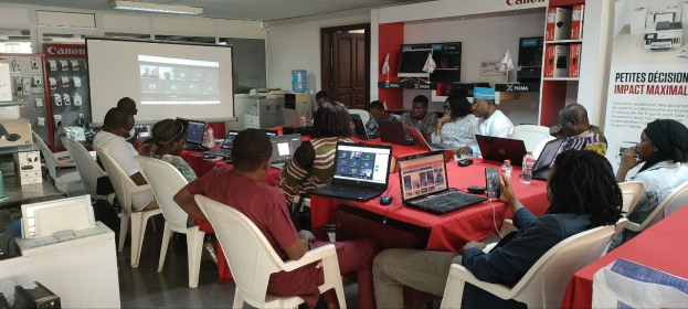 Gruppe von Menschen an einem Tisch mit Laptops, Mäusen und Flaschen während eines digitalen Marketing-Workshops, mit einem Banner, einem Bildschirm und Deckenleuchten im Raum.