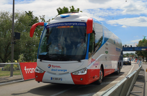 Ein roter und weißer Bus fährt auf einer Straße neben einem Zaun, mit Passagieren im Inneren, neben einem Geländer, Bäumen, Polen, Verkehrszeichen, einer Brücke und einem bewölkten Himmel.