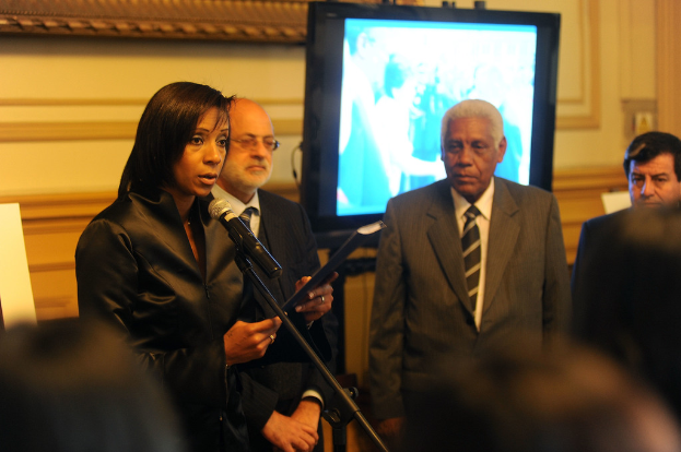 Eine Gruppe von Menschen drinnen mit einer Frau im Vordergrund vor einem Mikrofon und einem Fernseher und einer Wand im Hintergrund.